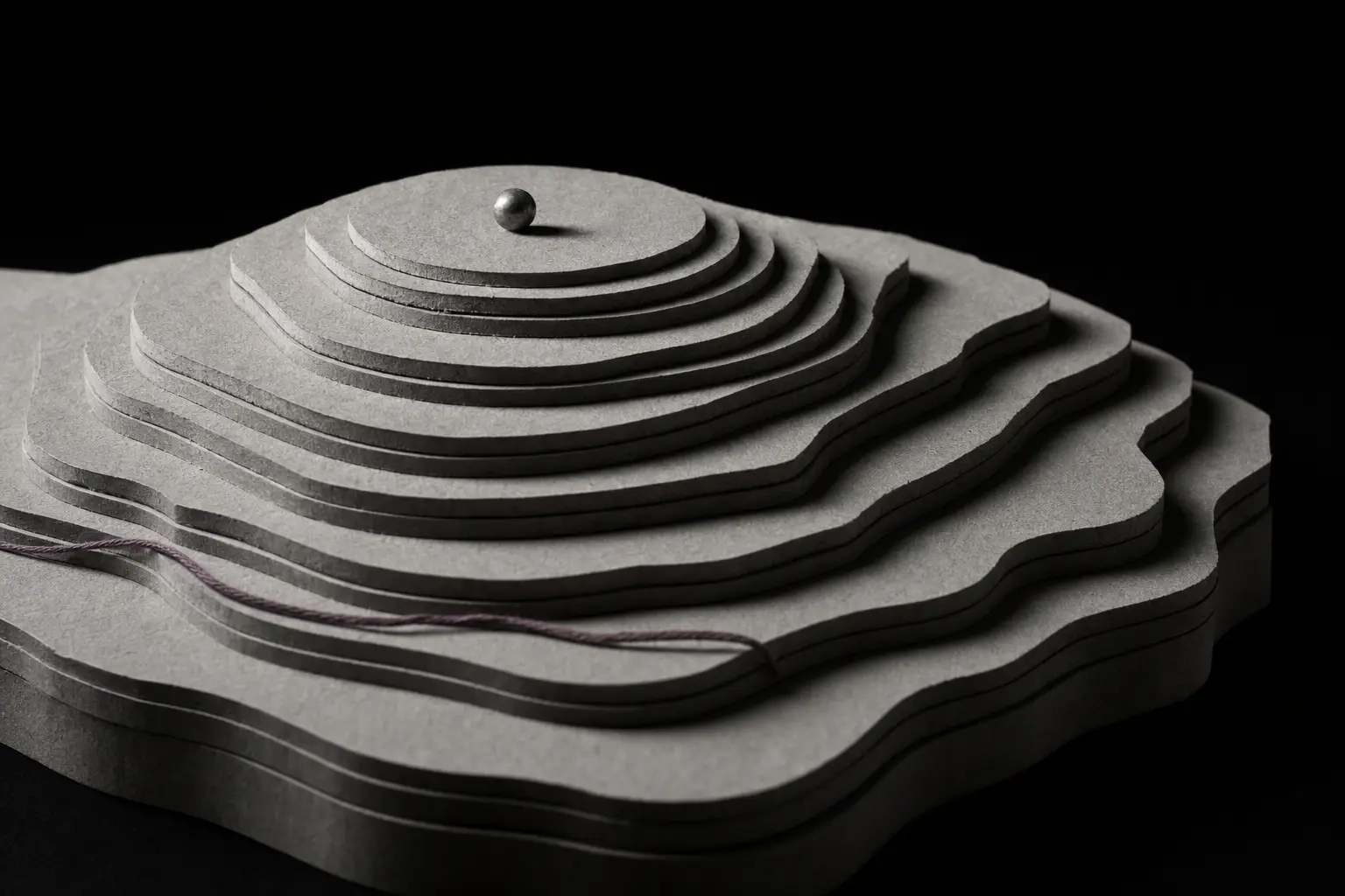 A relief model cut from many stacked layers of grey board, its terraces nesting into a broad summit. A single small steel sphere rests on the highest terrace, casting a small precise shadow. A plum thread lies along a lower terrace, far from the sphere.