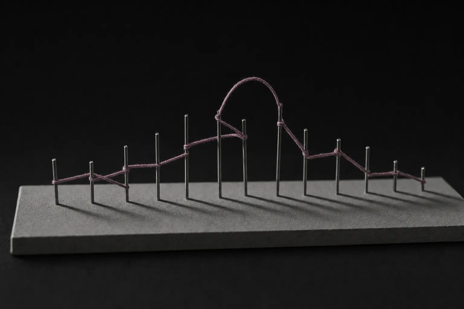 A row of slender steel pins standing at irregular heights on a grey board. A single continuous plum thread is wound between them, looping high above the row near the middle before returning to the line. Each pin casts a long hard shadow.