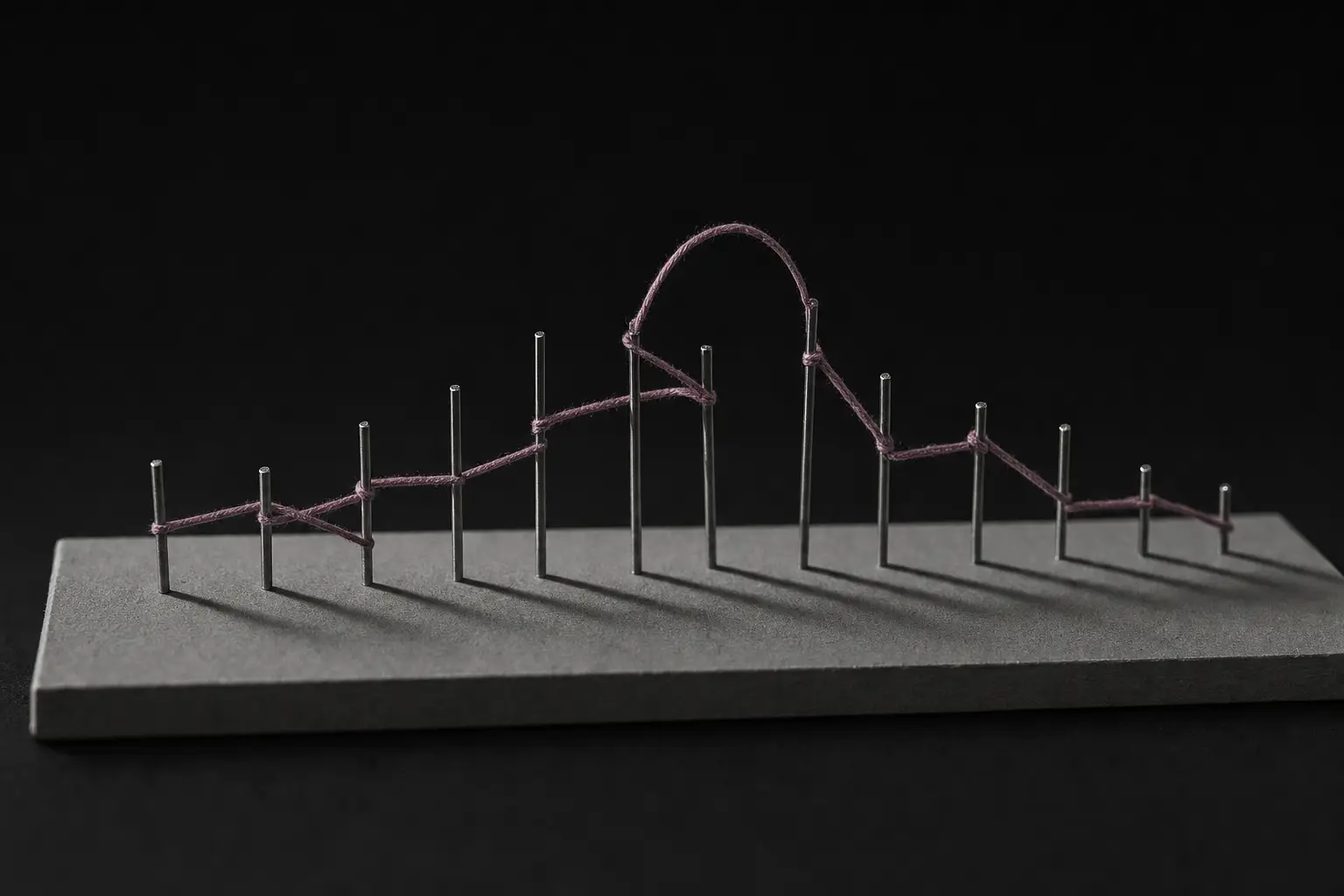A row of slender steel pins standing at irregular heights on a grey board. A single continuous plum thread is wound between them, looping high above the row near the middle before returning to the line. Each pin casts a long hard shadow.