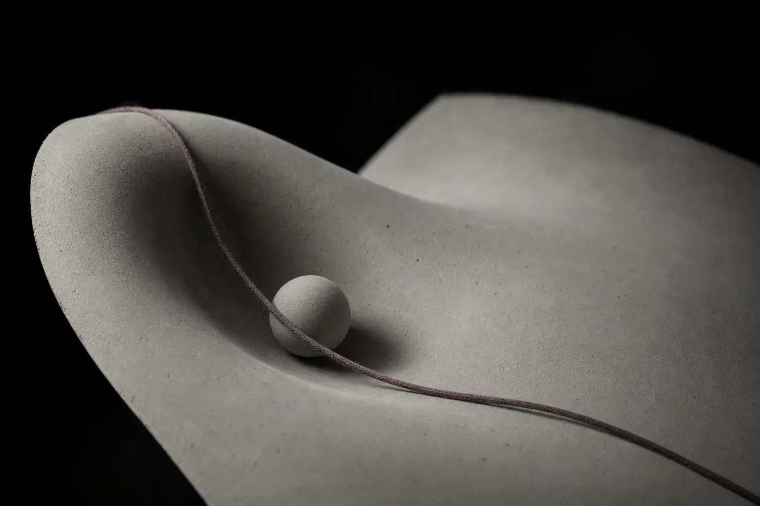 A smooth curved plaster surface, pale grey and matte, with one shallow depression worn into it. A matte grey sphere rests in the depression, held by the curvature. A plum thread is laid across the plaster along the line of steepest slope.