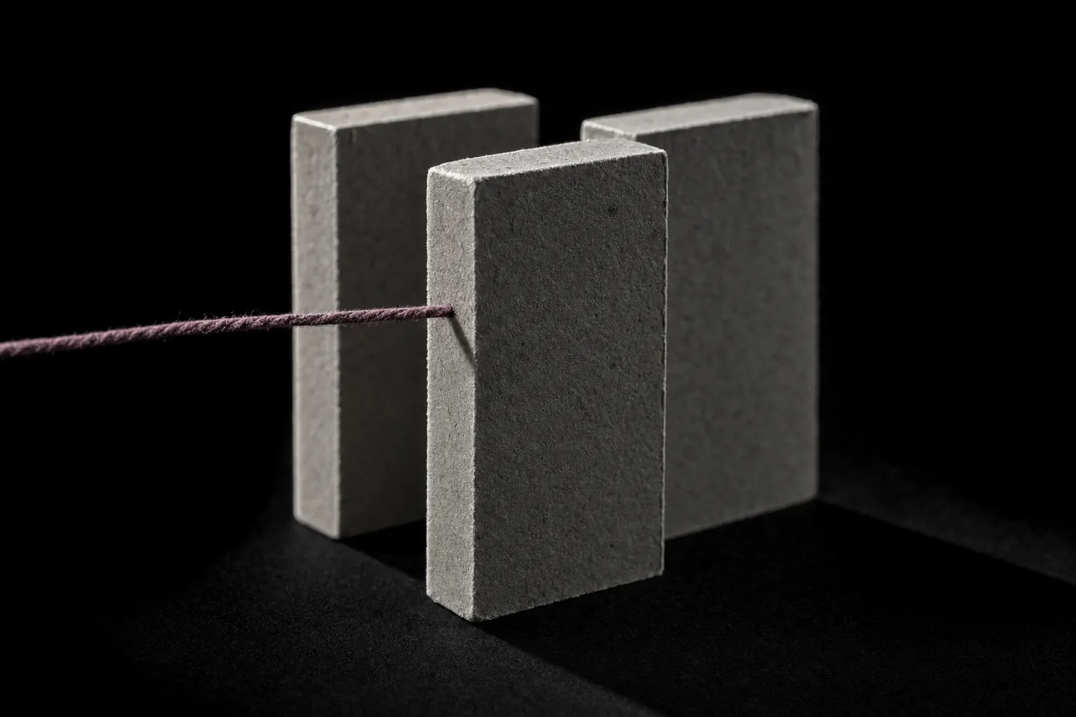 Three identical matte grey blocks standing in a close row against black, the middle one pulled forward. A single plum thread enters from outside the frame and touches only the forward block.