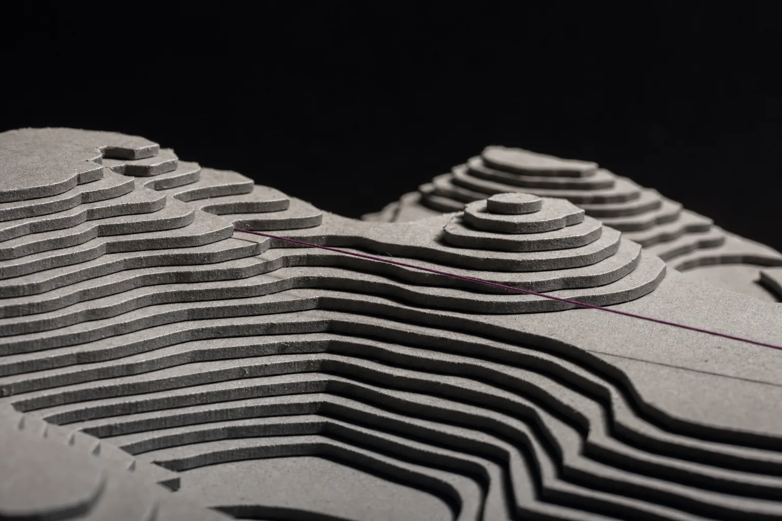 A wide relief model of terrain built from many stacked layers of cut grey board, forming nested terraces. A single taut plum thread is stretched low across it, casting one thin shadow. The model recedes into darkness at the back.